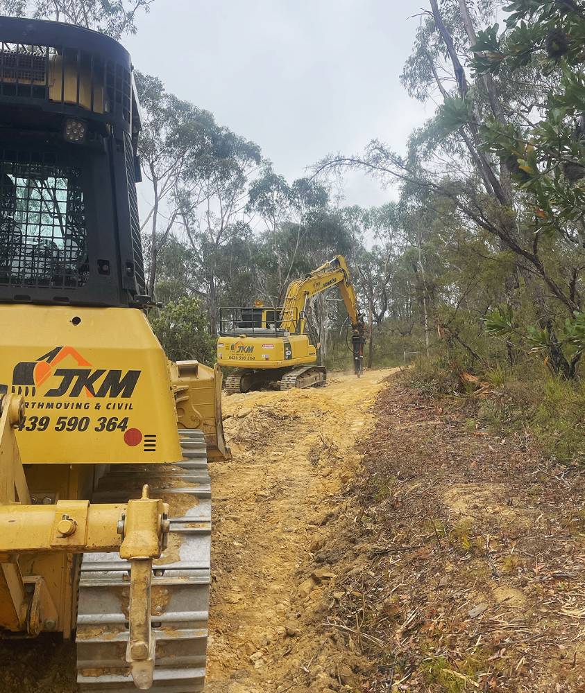 Yellow construction machinery, including a tracked excavator and a bulldozer with contact details visible, operate on a dirt path surrounded by trees in a forested area. Overcast sky in the background.