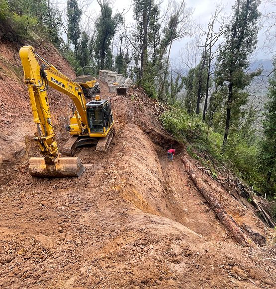 A yellow excavator is parked on a muddy, terraced hillside surrounded by trees. A person with a red umbrella stands near the excavator in an excavated section, indicating the steep terrain and ongoing construction work.