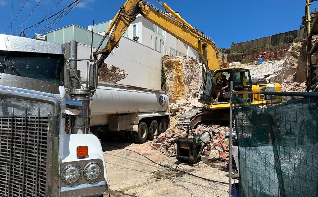 A construction site with a yellow excavator loading debris into a large white truck. Rubble and broken concrete are scattered around. A worker is operating the excavator. The sky is clear and blue, with a modern building in the background.