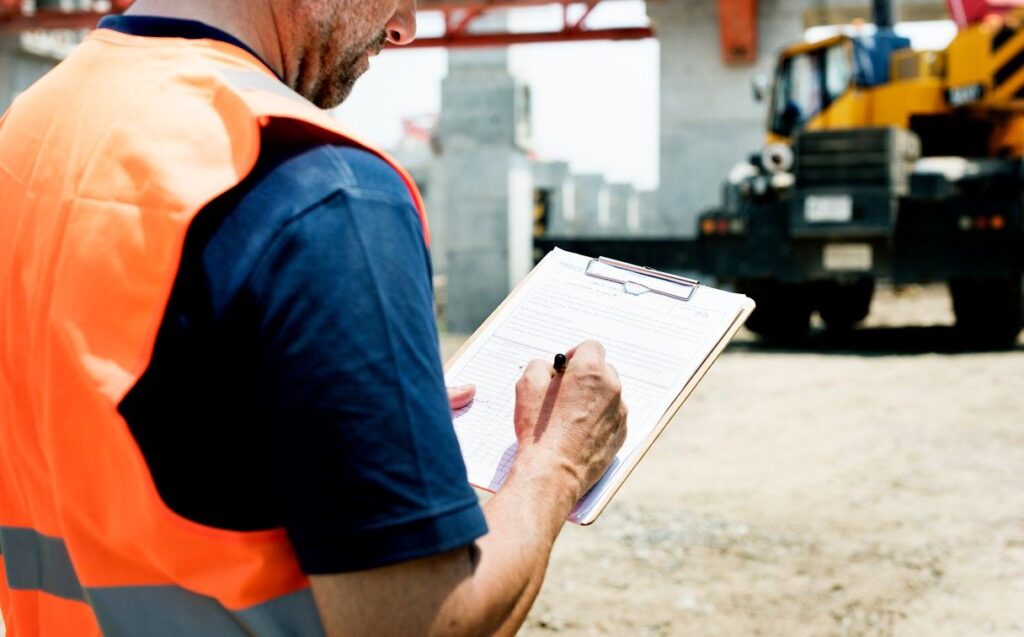 A construction worker wearing a bright orange safety vest is writing on a clipboard at a construction site. In the background, there's heavy machinery and unfinished structures under a clear sky.