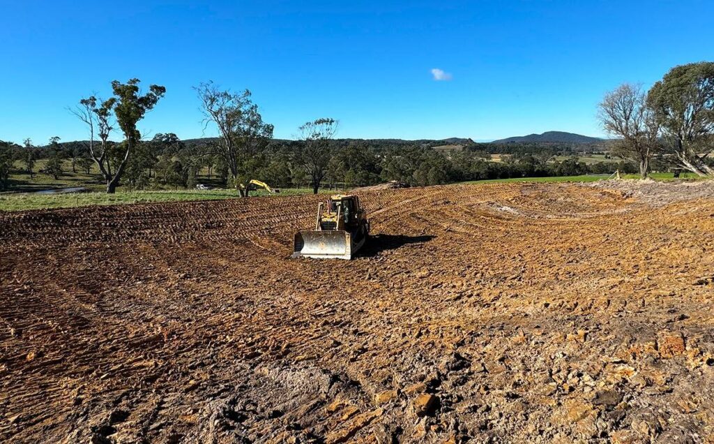 A bulldozer works on a large dirt area surrounded by grassy fields and scattered trees under a clear blue sky. Hills are visible in the distance.