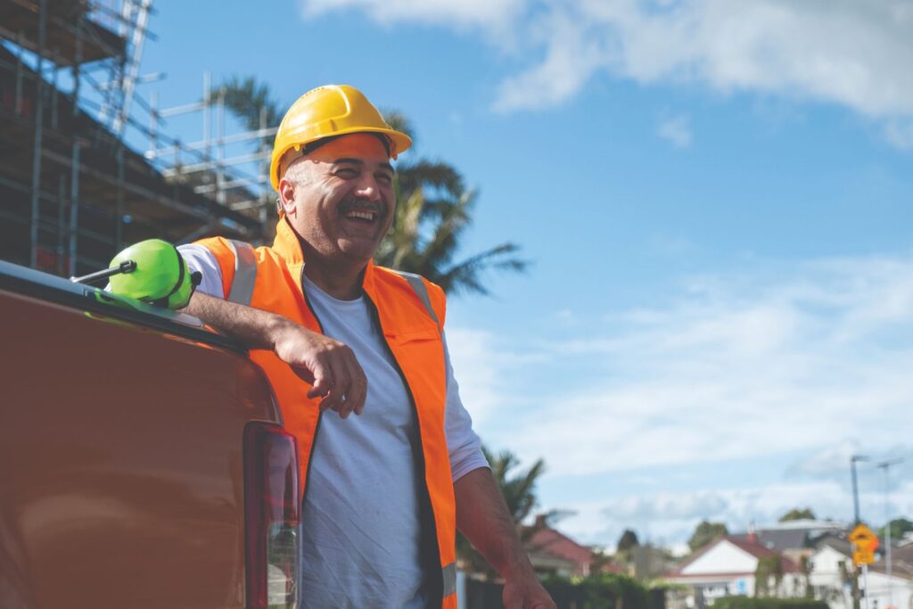 A construction worker in an orange safety vest and yellow hard hat leans against a truck, smiling. A pair of safety gloves rests beside him. The background shows a building under construction and a clear blue sky with some clouds.