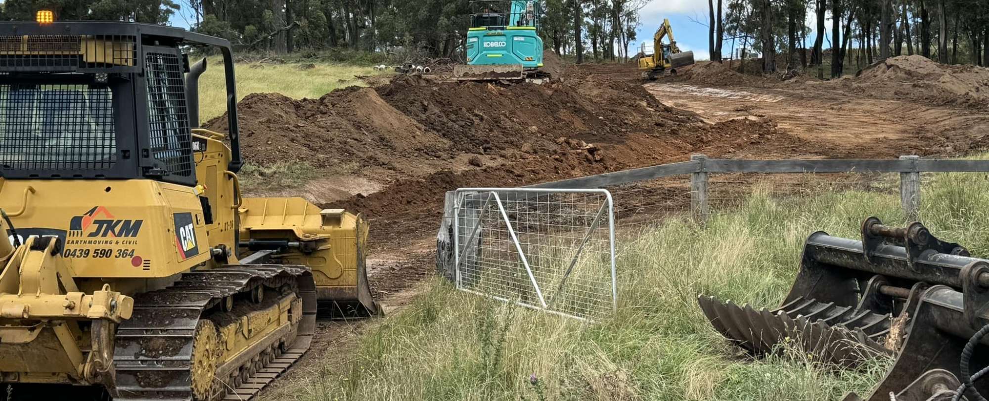 Construction site with heavy machinery, including a yellow bulldozer and an excavator on a dirt road surrounded by grassy fields and trees. An open metal gate and a pile of large machine attachments are visible in the foreground.