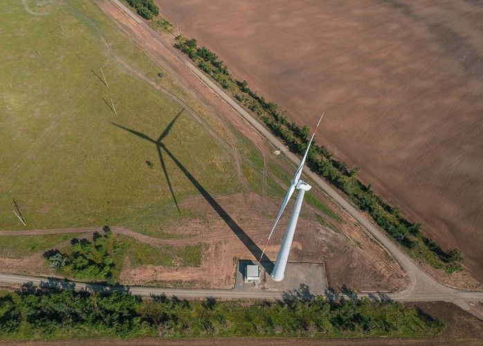 Aerial view of a wind turbine casting a long shadow on a grassy field. A dirt road runs alongside the turbine, with trees lining the path. Adjacent is a plowed farmland section, contrasting with the green landscape.