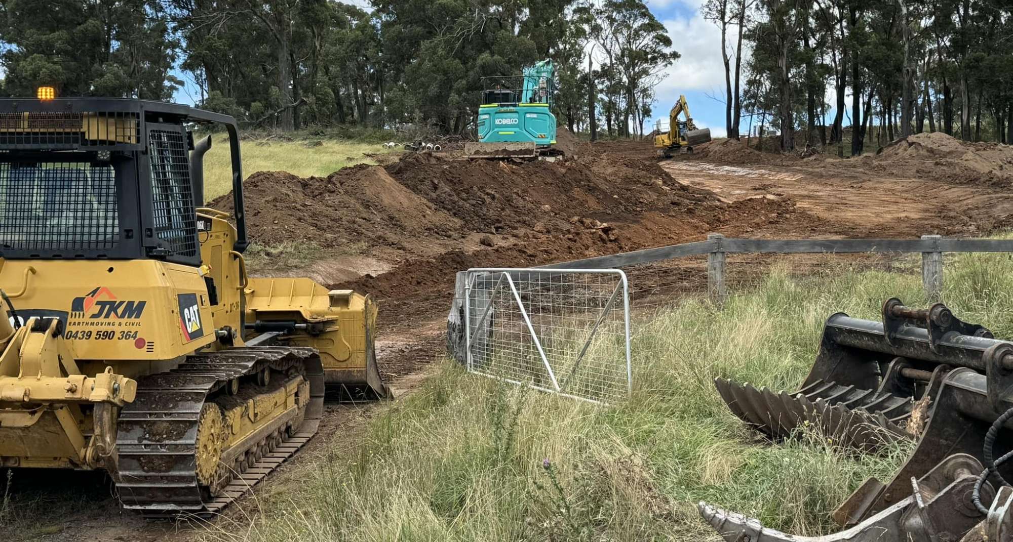 Construction site with two bulldozers, one green and one yellow, on a dirt road surrounded by grassy fields and trees. A metal gate is open by the path, and a pile of mechanical equipment lies on the right side.