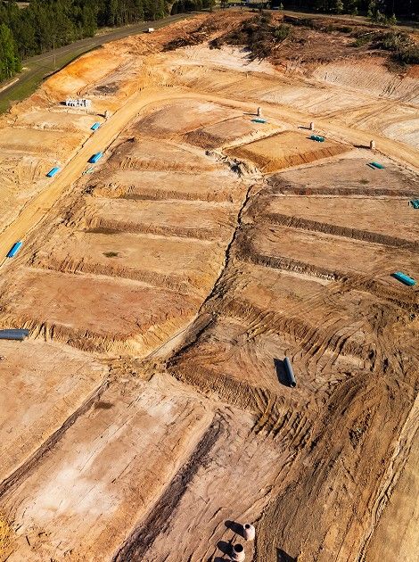 Aerial view of a construction site with leveled plots of land. The area is divided into rectangular sections with exposed soil and scattered green pipes. Surrounding the site are trees and patches of grass.