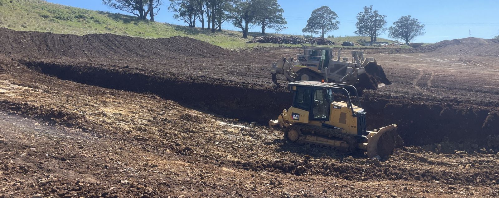 Construction site with two bulldozers moving earth in a large, open area. The terrain is uneven, with mounds of soil and clear blue sky above. Trees are visible in the background.
