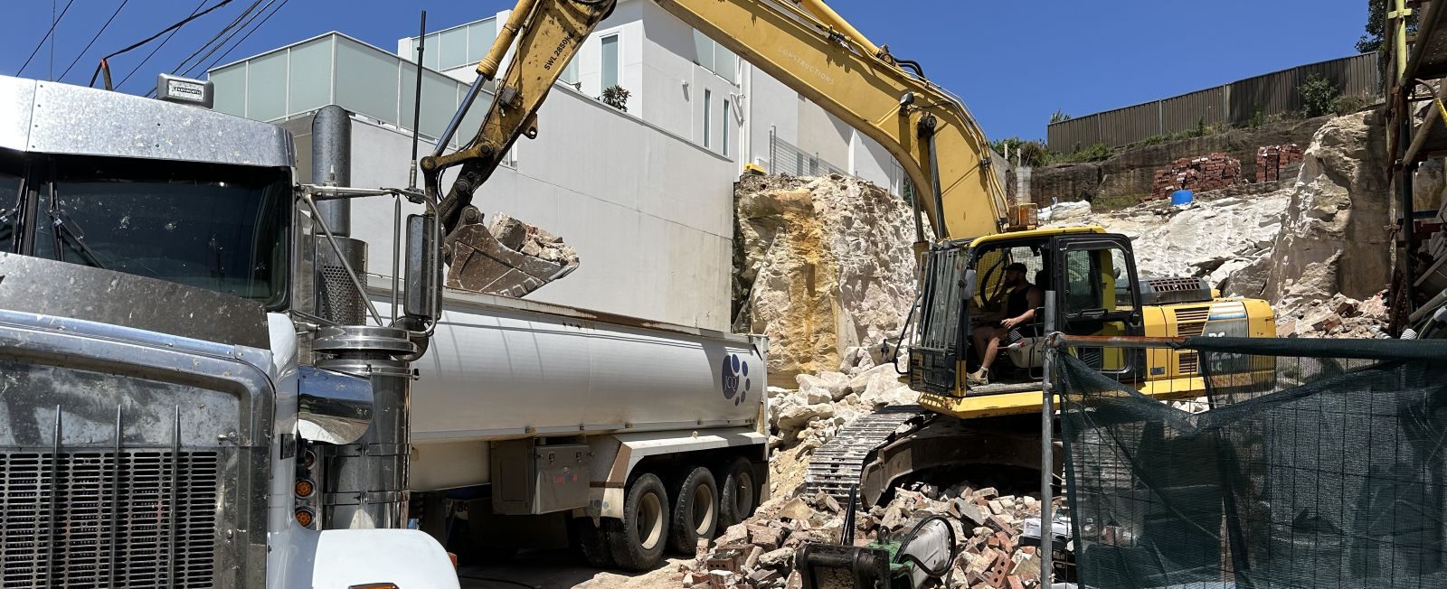 A construction site with a yellow excavator loading debris into a large white dump truck. The area has rocky terrain and partially demolished structures in the background, under a clear blue sky. A green fence is visible in the foreground.