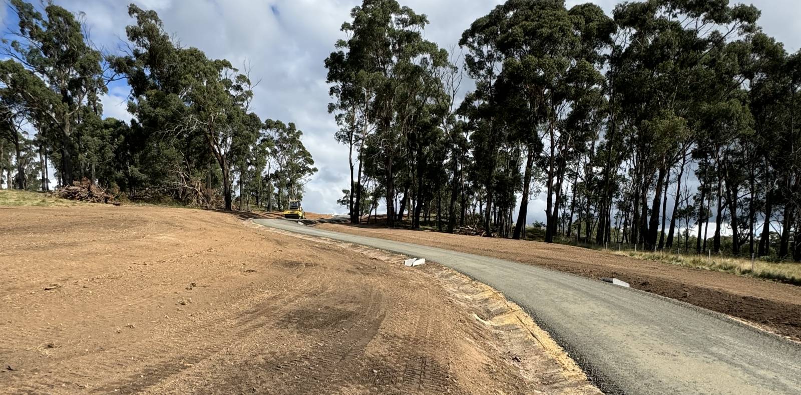 A freshly paved road curves through a landscape bordered by tall trees on the right. The left side shows cleared, earthy terrain. The sky is mostly cloudy, with patches of blue visible.