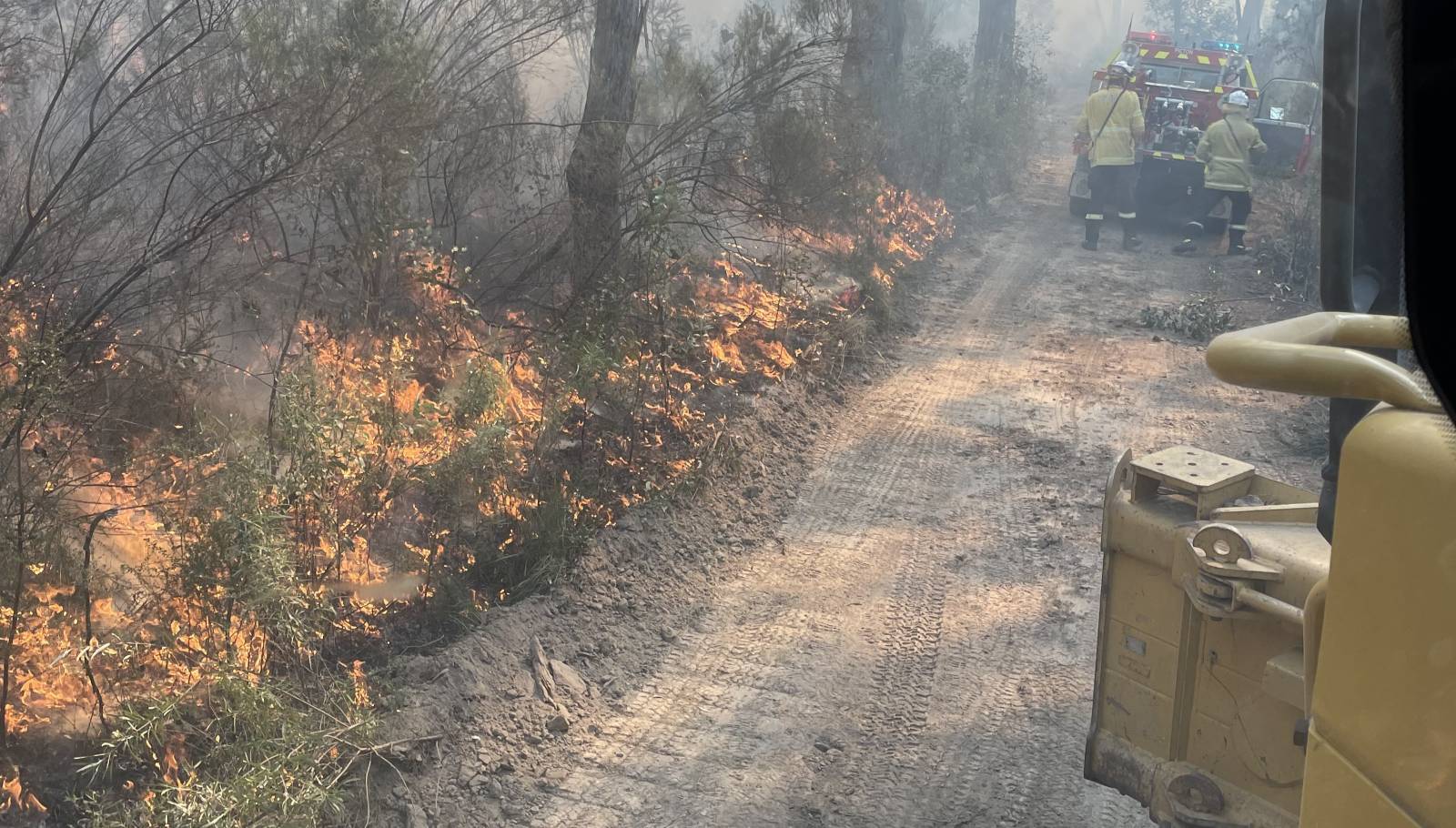 A dirt road bordered by a forest is burning with flames visible on the left. Smoke fills the air, and firefighters in yellow gear are gathered beside a fire truck on the right, working to control the wildfire.