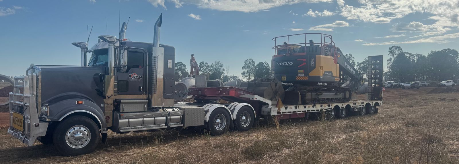 A heavy-duty truck is parked on a grassy area with a flatbed trailer carrying a large Volvo excavator. The sky is partially cloudy, and trees are visible in the background.