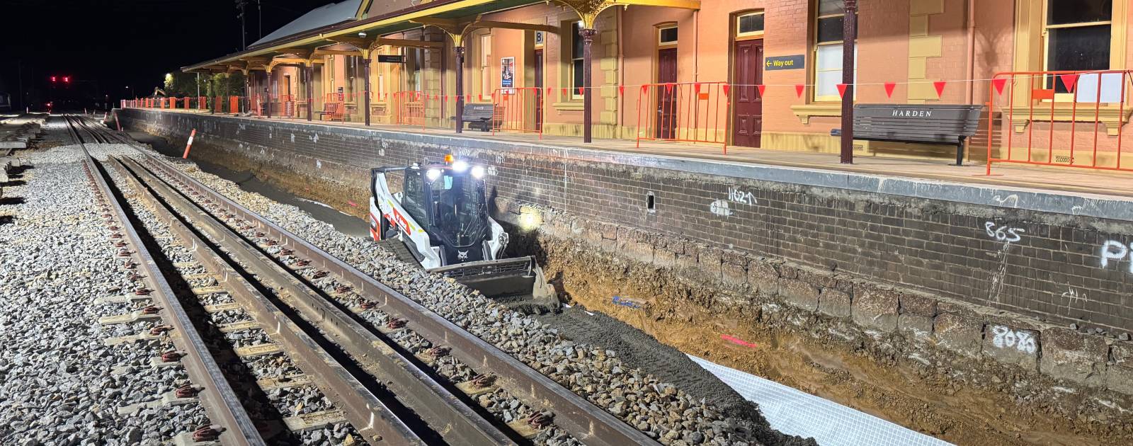 A small construction vehicle works on railway track maintenance at night next to an illuminated train station platform. The platform has a covered waiting area and benches, with red barriers along the edge.
